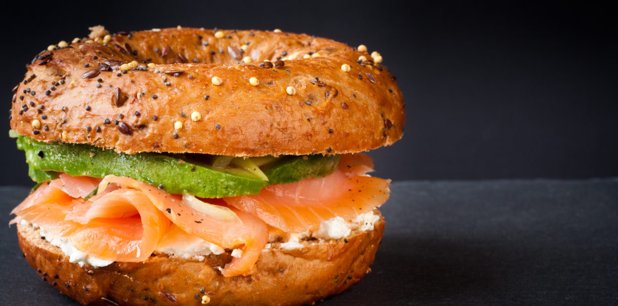 Healthy freshly baked bagel filled with smoked salmon lox and topped with avocado. Served on a gray slate table against a dark background.